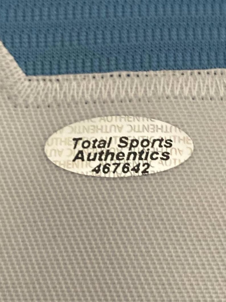 May include: Close-up of a light gray fabric with a white stitched edge and a blue fabric in the background. An oval sticker with the text "Total Sports Authentics" and the number "467642" is visible.
