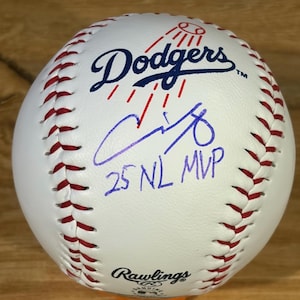 May include: A white leather baseball with red stitching featuring the blue Dodgers logo, a red graphic of a baseball, a purple ink signature, and the handwritten text 25 NL MVP. The Rawlings brand logo is printed in black at the bottom of the ball.
