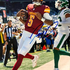 May include: A signed photograph of a football player wearing a burgundy Commanders jersey with the number 5 in yellow, white trousers, and a helmet, running on a pitch while holding a football, with a blue ink autograph across the centre.