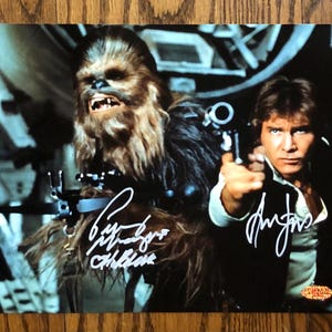 May include: A color photograph featuring Chewbacca and Han Solo from Star Wars. Both characters are in the foreground, with Han Solo holding a blaster. The image is signed by the actors. The background is a spaceship interior.