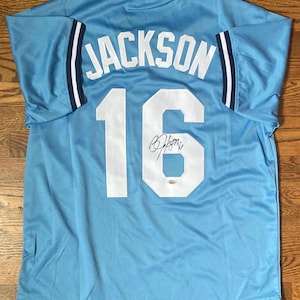 May include: Light blue baseball jersey with the name "JACKSON" and the number "16" in white on the back. The jersey has a signature below the number. The sleeves have navy blue and white stripes.