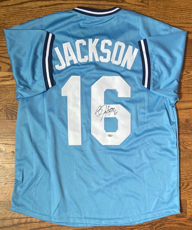 May include: Light blue baseball jersey with the name "JACKSON" and the number "16" in white on the back. The jersey has a signature below the number. The sleeves have navy blue and white stripes.