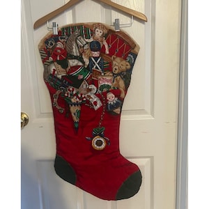 May include: A red fabric Christmas stocking featuring a printed collage of vintage toys including a rocking horse, a teddy bear, a doll, a toy soldier, and a train. A small clock charm hangs from the stocking, which displays the text Merry Christmas.