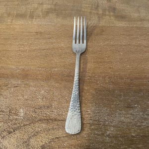 以下が含まれることがあります： 4本歯の銀色のフォーク。ハンドルはテクスチャ加工されたハンマー加工のデザインです。フォークは、自然な木目を持つ木製の表面の中央に配置されています。