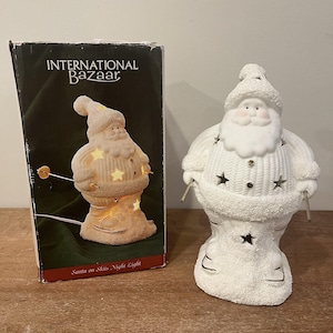 May include: A white ceramic Santa Claus night light with star-shaped cutouts. The figure is wearing a knitted hat and sweater, and is standing on skis. The box next to it reads "INTERNATIONAL BAZAAR" and "Santa on Skis Night Light."
