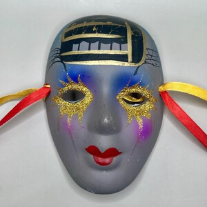 May include: A decorative ceramic mask with a gray face, red lips, and gold glitter around the eyes. The mask has blue and gold accents on the forehead and red and yellow ribbons attached to the sides.