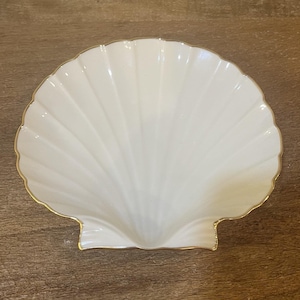 May include: A white, shell-shaped decorative dish with a gold trim. The dish has a ribbed texture and a scalloped edge. The dish is made of ceramic or porcelain and is likely used for serving or display.