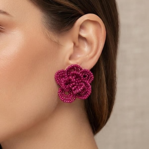 Pode incluir: Um brinco de tarracha em forma de flor na cor rosa fúcsia, feito com pequenas missangas densamente agrupadas. O brinco apresenta um design de pétalas circulares em camadas que cria uma aparência texturizada e tridimensional.
