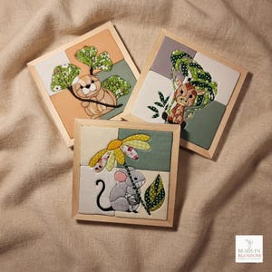 Pode incluir: Três peças de arte de parede quadradas em tecido com moldura de madeira e aplicações de animais. Uma mostra uma lontra castanha com folhas verdes, uma uma girafa com folhagem verde e uma um rato cinzento a segurar uma flor amarela e branca.