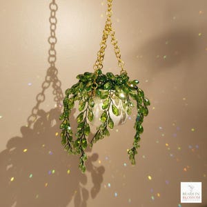 Pode incluir: Um ornamento de planta suspensa com contas de cristal verde facetadas em forma de folhas, suspenso por uma corrente de metal dourado. As contas estão dispostas em vinhas em cascata em torno de um pequeno vaso branco. O logótipo BEADS IN BLOSSOM é visível.