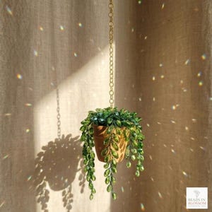 Op de afbeelding: Een hangende terracotta plantenpot met een trapsgewijze groene plant, opgehangen aan een gouden ketting. De plant heeft groene, bladvormige versieringen. De achtergrond is een beige gordijn met lichtreflecties. De pot is gelabeld met de tekst "BEADS IN BLOSSOM".