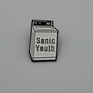 May include: A white and black enamel pin in the shape of a washing machine. The pin features the text "Sonic Youth" in a bold, sans-serif font. The pin has a black border and is set against a gray background.