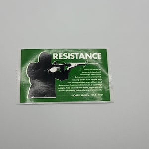 May include: A green sticker with the word "RESISTANCE" in white text. A black silhouette of a person holding a rifle is on the left. Text from Bobby Sands is included.
