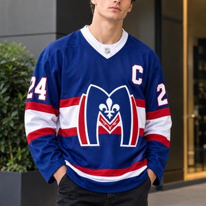 May include: A blue hockey jersey featuring a white V-neck collar, a large stylized letter M with a fleur-de-lis emblem on the chest, the number 24 on the sleeve, the number 2 on the other sleeve, and a white C on the upper chest area.