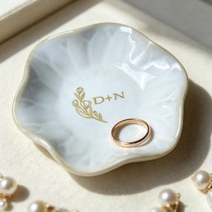 May include: A white ceramic ring dish with a scalloped edge and a glossy finish. The center features a gold-colored botanical illustration and the text D+N. A plain gold-colored metal band rests inside the dish, which has a smooth, curved surface.