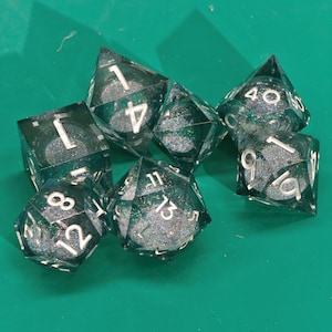 Puede incluir: Un conjunto de siete dados poliédricos transparentes llenos de purpurina con numeración blanca. Los dados tienen varias formas, incluyendo un cubo, un tetraedro y un dado de 20 caras. Los dados están colocados sobre una superficie verde.