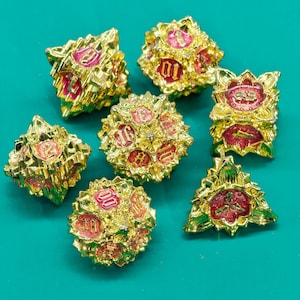 May include: A collection of seven gold-colored polyhedral dice with red and pink floral accents. Each die features a different number, with the numbers in a pink font. The dice are arranged on a teal surface.