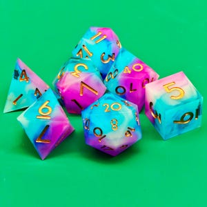 Op de afbeelding: Een set van zeven polyhedrale dobbelstenen in verschillende vormen, waaronder een d4, d6, d8, d10, d12, d20 en een percentieldobbelsteen. De dobbelstenen zijn gemarmerd met blauwe, paarse en witte hars, met goudkleurige cijfers.