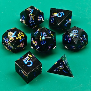 Puede incluir: Conjunto de siete dados poliédricos negros con diseños coloridos y brillantes. Cada dado presenta una forma y un número de caras diferentes, con números y símbolos azules, amarillos y rosas. Dados diseñados para juegos de rol de mesa.