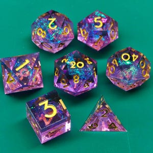 May include: A set of seven polyhedral dice in various shapes, including a d4, d6, d8, d10, d12, and d20. The dice are transparent purple with gold-colored numbers and iridescent inclusions. The dice are for tabletop games.