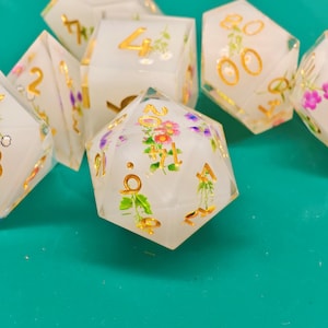 Puede incluir: Un conjunto de dados poliédricos con un diseño floral. Los dados son blancos con números dorados y detalles florales en rosa, morado y verde. Los dados están hechos de un material de resina transparente.