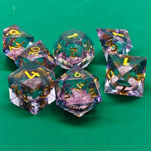 Könnte beinhalten: Ein Satz von sieben polyedrischen Würfeln für Tabletop-Spiele. Die transparenten Würfel haben ein türkis-violettes Glitzer-Interieur und goldene Zahlen. Die Würfel liegen auf einer grünen Oberfläche.