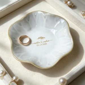 Könnte beinhalten: Eine weiße Keramikschale für Ringe mit gewelltem Rand und Goldrand. In der Mitte befindet sich ein goldener Aufdruck mit einem Blattzweig, den Namen Martin und Sophia, getrennt durch ein Herz, und dem Datum 06.24.2026. Ein goldener Ring liegt darin.