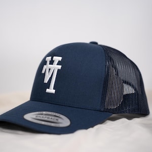May include: Navy blue trucker hat with a white embroidered "VT" logo on the front. The hat features a curved brim and a mesh back for breathability. The snapback closure allows for an adjustable fit.