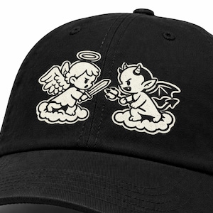 Op de afbeelding: Zwarte baseballcap met een cartoon engel en duivel ontwerp. De engel, met een halo en zwaard, staat tegenover de duivel, die hoorns en een drietand heeft. Beide personages staan op wolken.