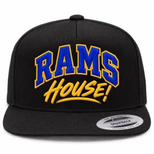 May include: A black snapback cap featuring the embroidered text RAMS in blue with a yellow outline, and the word HOUSE! in yellow script below it. A circular silver and black sticker is affixed to the flat brim, reading THE AUTHENTIC YUPOONG SNAPBACK.