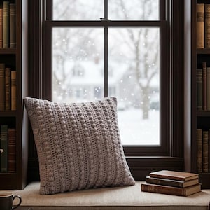 May include: A square, light gray crochet pillow featuring a textured bobble stitch pattern and a side panel accented with a vertical row of small, round buttons. The pillow is positioned on a window seat in front of a window with a snowy view.