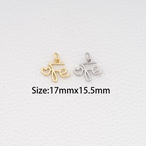 May include: Two metal pendants, one gold-toned and one silver-toned, each shaped like the word "Fe" with a heart. The pendants are approximately 17mm x 15.5mm. The word "Fe" is written in a cursive font.
