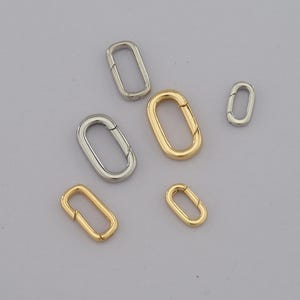 May include: An assortment of oval-shaped metal clasps in silver and gold tones. The clasps vary in size, with a mix of open and closed designs. The background is a solid, neutral color, highlighting the metallic sheen of the clasps.