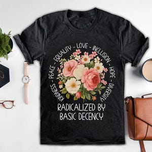 Könnte beinhalten: Schwarzes T-Shirt mit einem Blumendesign aus rosa und weißen Blumen. Das Design ist von Wörtern wie "Gleichheit", "Liebe" und "Inklusion" umgeben. Der Text "Radikalisiert durch grundlegende Anständigkeit" steht darunter.