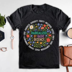 Pode incluir: Camiseta cinza escuro com um design floral que envolve as palavras "RADICALIZED BY BASIC DECENCY". O design inclui uma variedade de flores coloridas e as palavras "Diversidade, Equidade, Inclusão, Justiça, Unidade, Cuidado, Compaixão, Paz, Mudança, Comunidade, Amor, Bondade."