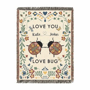 May include: A cream-colored woven throw blanket featuring two ladybugs in the center with the text LOVE YOU, KATE, JOHN, and LOVE BUG. The design includes a floral border with green vines, red, yellow, and blue flowers, butterflies, and dragonflies.