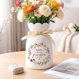 May include: A white ceramic vase with a floral arrangement of orange, pink, yellow, and white flowers. The vase has a gold ribbon and a floral wreath design with the words "mom grandma" and the years "EST. 1996" and "EST. 2024".