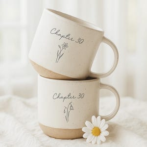May include: Two stacked ceramic mugs with a cream-colored upper half and a tan base. Each mug has the text "Chapter 30" and a floral illustration. A white daisy with a yellow center rests near the bottom mug.
