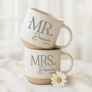 May include: Two stacked off-white ceramic mugs with a beige base. The top mug reads "MR. Brown 15.09.25" in gray lettering. The bottom mug reads "MRS. Brown 15.09.25". A white daisy with a yellow center rests near the bottom mug.