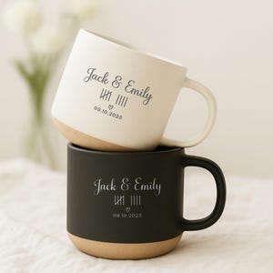 May include: Two stacked ceramic mugs. The top mug is white with a tan base, and the bottom mug is black with a tan base. Both mugs have the names "Jack & Emily" and a date printed on them.