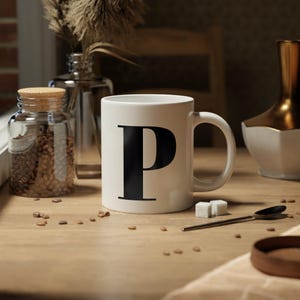 Può includere: Tazza in ceramica bianca con una grande lettera nera "P" sul davanti. La tazza è su una superficie di legno con chicchi di caffè, zollette di zucchero e un cucchiaio. Un barattolo di vetro con coperchio in sughero e un vaso decorativo sono sullo sfondo.
