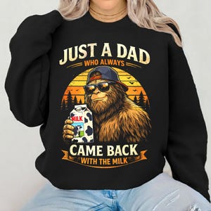 Może przedstawiać: Czarna bluza z grafiką Bigfoota w okularach przeciwsłonecznych i czapce z daszkiem, trzymającego karton mleka. Tekst brzmi "JUST A DAD WHO ALWAYS CAME BACK WITH THE MILK" w projekcie zachodu słońca.