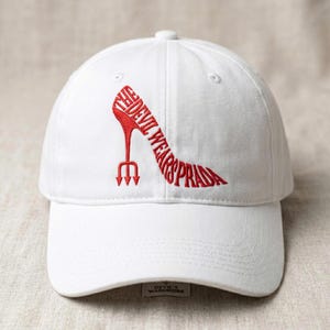 May include: White baseball cap with a red embroidered design of a high-heeled shoe and the text "THE DEVIL WEARS PRADA." The cap has a curved brim and a small tag that reads "DEVIL'S WARDROBE."