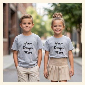 May include: Two children wearing heather gray short-sleeved t-shirts with the black text Your Design Here printed on the chest. One child wears beige cargo shorts, and the other wears a beige pleated skirt. The shirts have a crew neckline.
