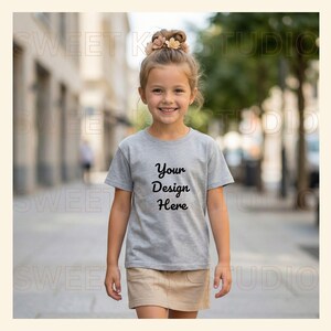 May include: A young girl wearing a gray t-shirt and a tan skirt walks down a city street. The t-shirt has the text "Your Design Here" in black script. The girl has her hair up and is smiling.