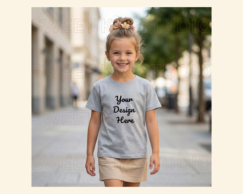 May include: A young girl wearing a gray t-shirt and a tan skirt walks down a city street. The t-shirt has the text "Your Design Here" in black script. The girl has her hair up and is smiling.