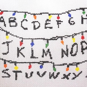 May include: Cross-stitch design showing the alphabet from A to Z in black. The letters are arranged in three rows, with colourful string lights above and below each row. The background is white.