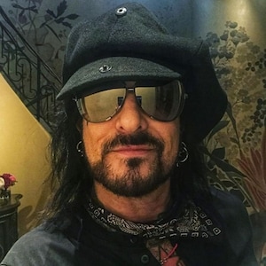 May include: A man with long black hair and a beard is wearing a black newsboy cap, sunglasses, a black bandana, and a black shirt. He is smiling at the camera.