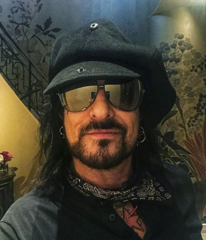 May include: A man with long black hair and a beard is wearing a black newsboy cap, sunglasses, a black bandana, and a black shirt. He is smiling at the camera.