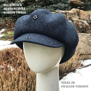 May include: A blue and black herringbone Harris Tweed newsboy cap with a smaller version label. The cap is on a white mannequin head.